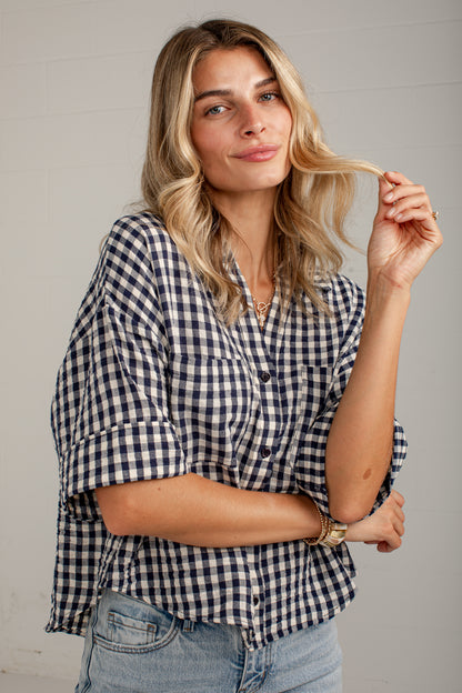 A woman wearing a navy and white gingham check button-down shirt with rolled sleeves, paired with light-wash blue jeans, standing against a light gray paneled wall.