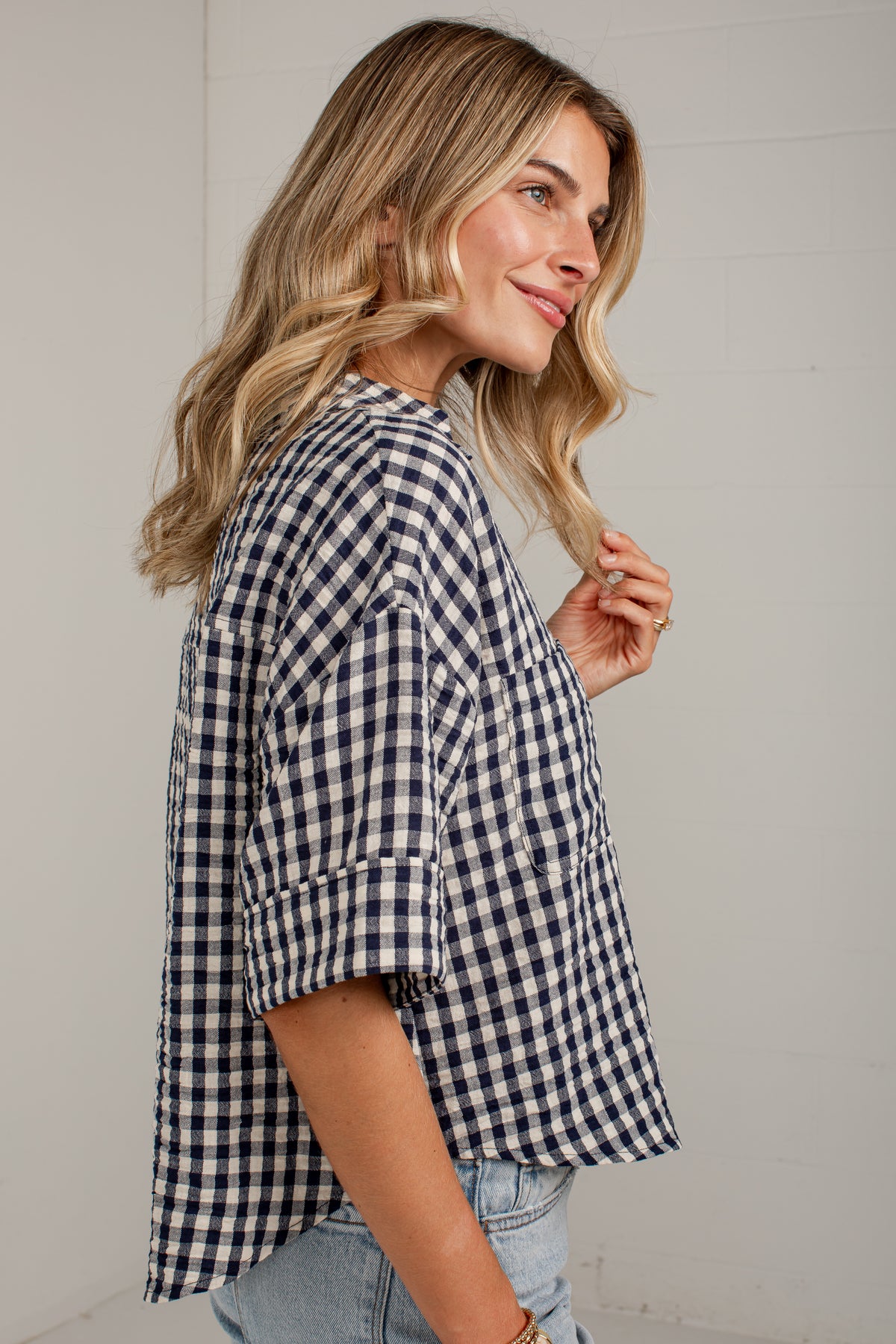 A woman wearing a navy and white gingham check short-sleeve button-up shirt with rolled cuffs, paired with light-wash jeans, shown from a side profile against a white paneled wall.