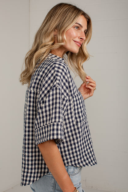 A woman wearing a navy and white gingham check short-sleeve button-up shirt with rolled cuffs, paired with light-wash jeans, shown from a side profile against a white paneled wall.