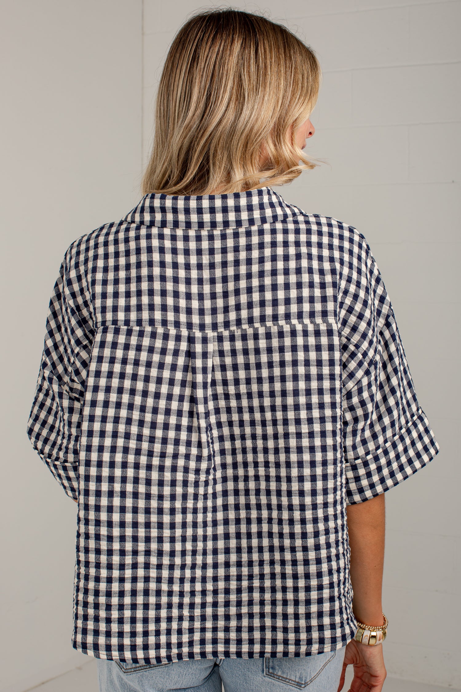 A woman viewed from behind wearing a navy and white gingham short-sleeve button-up shirt with a large back pocket, paired with light blue jeans.