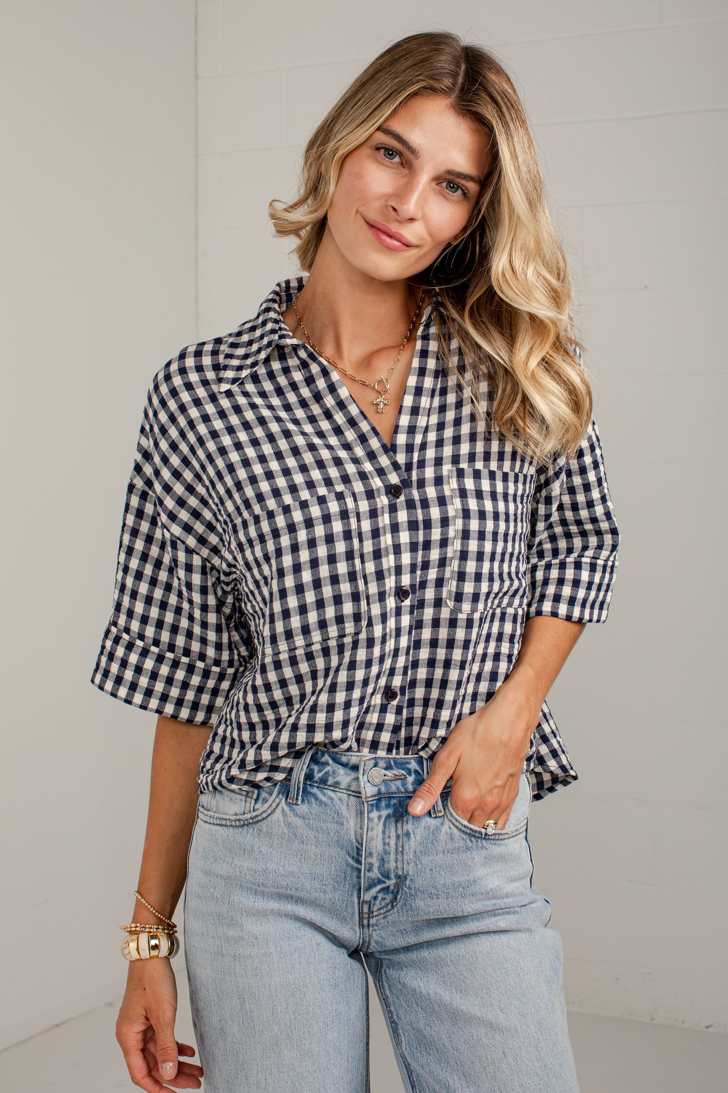 A woman wearing a navy and white gingham button-up shirt with rolled-up sleeves and light-wash blue jeans, standing against a light gray wall.