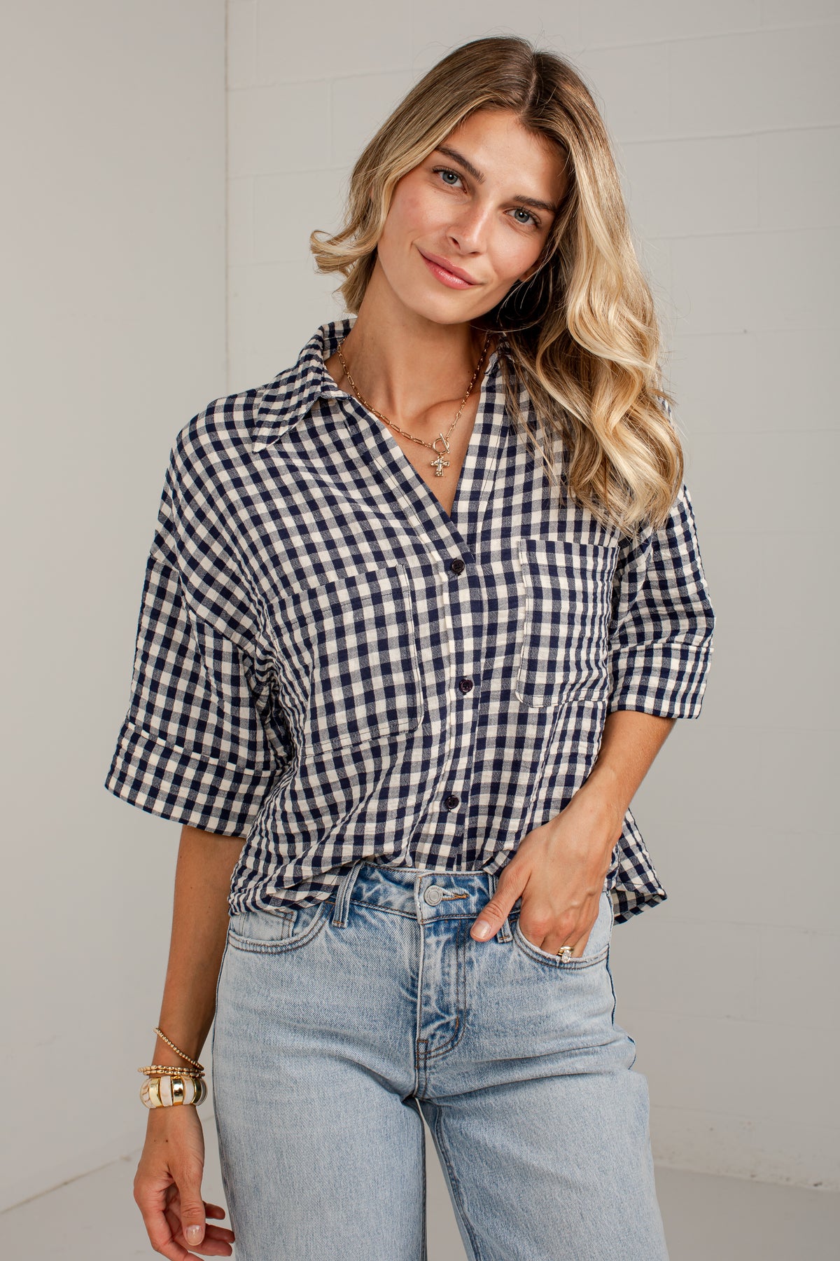 A woman wearing a navy and white gingham button-up shirt with rolled-up sleeves and light-wash blue jeans, standing against a light gray wall.
