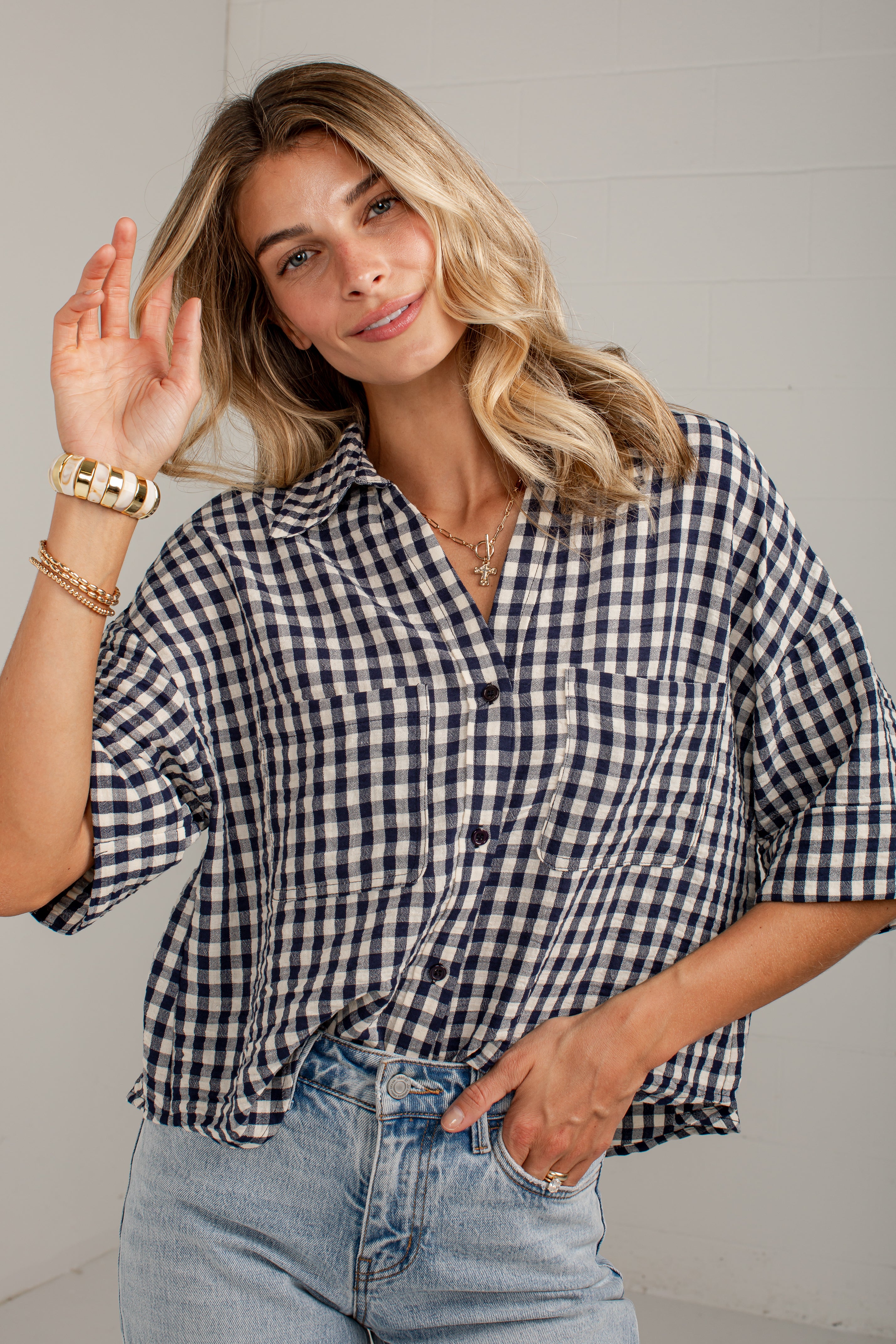 A woman wearing a navy and white gingham short-sleeve button-up shirt with light blue jeans, posing against a light gray paneled wall.