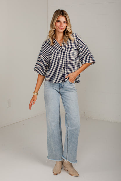 A woman modeling a black and white gingham short-sleeve button-up shirt with light-wash wide-leg jeans and beige ankle boots, standing in a bright white studio.