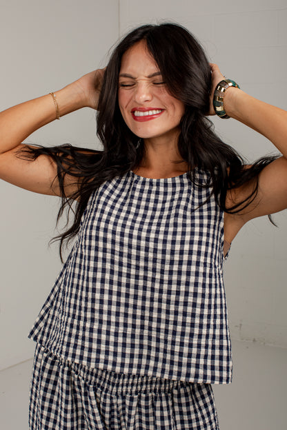 A woman with long dark hair wearing a navy and white gingham check sleeveless top and matching bottoms, posing with her hands behind her head against a plain light gray wall.