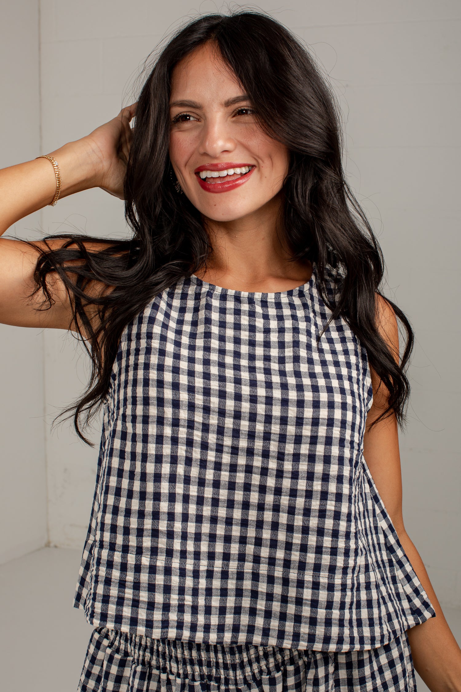 A woman with long dark hair smiling while wearing a navy and white gingham check sleeveless top and matching bottoms, posed against a light gray wall.