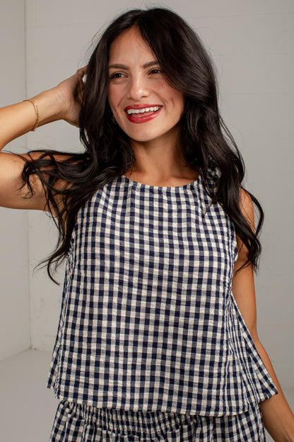 A woman with long dark hair smiling while wearing a navy and white gingham check sleeveless top and matching bottoms, posed against a light gray wall.