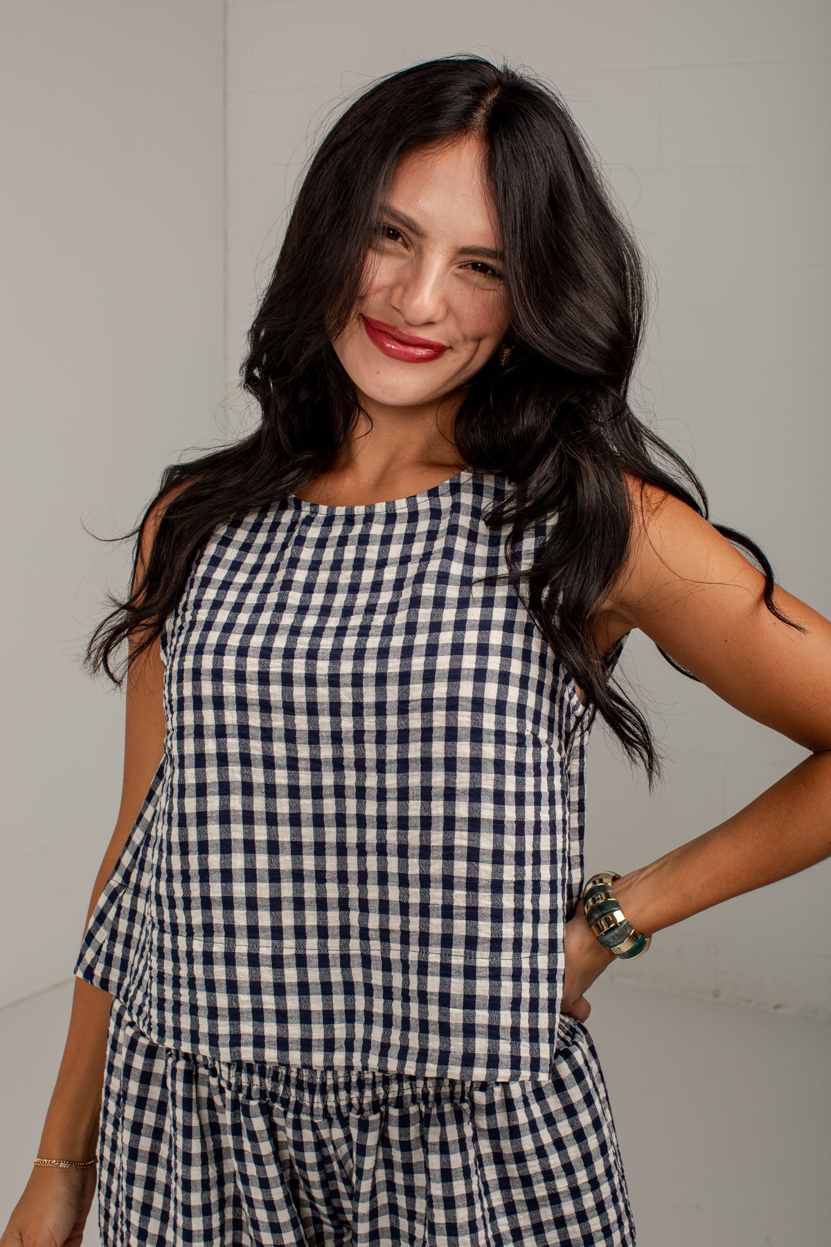A woman with long dark hair wearing a sleeveless navy and white gingham check top, posing against a plain light gray wall.