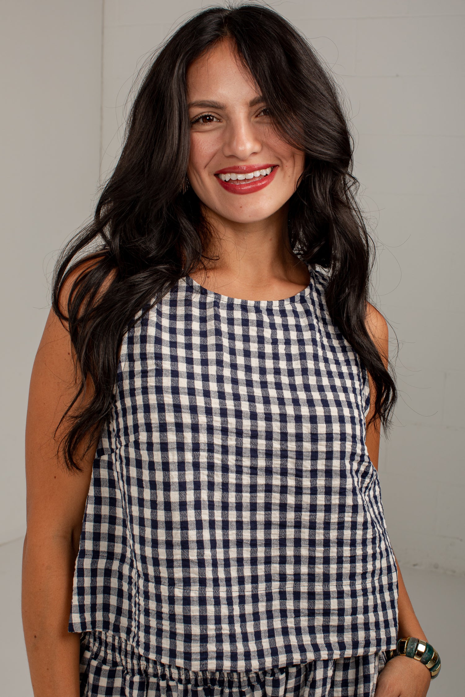 A smiling woman with long dark hair wearing a sleeveless navy and white gingham check top, photographed against a plain light gray wall.