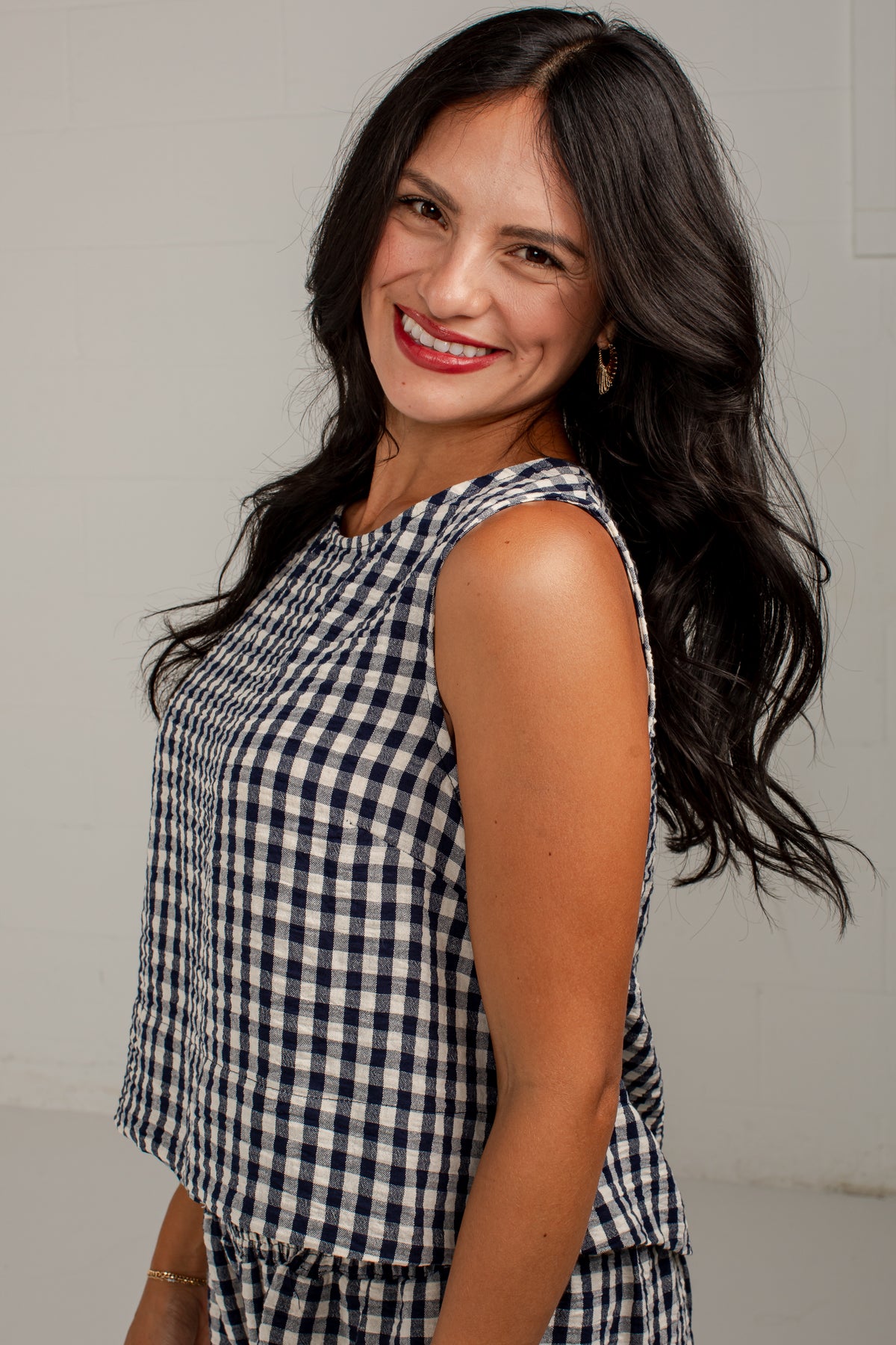 A smiling woman with long dark hair wearing a sleeveless navy and white gingham check top, photographed against a plain light gray background.