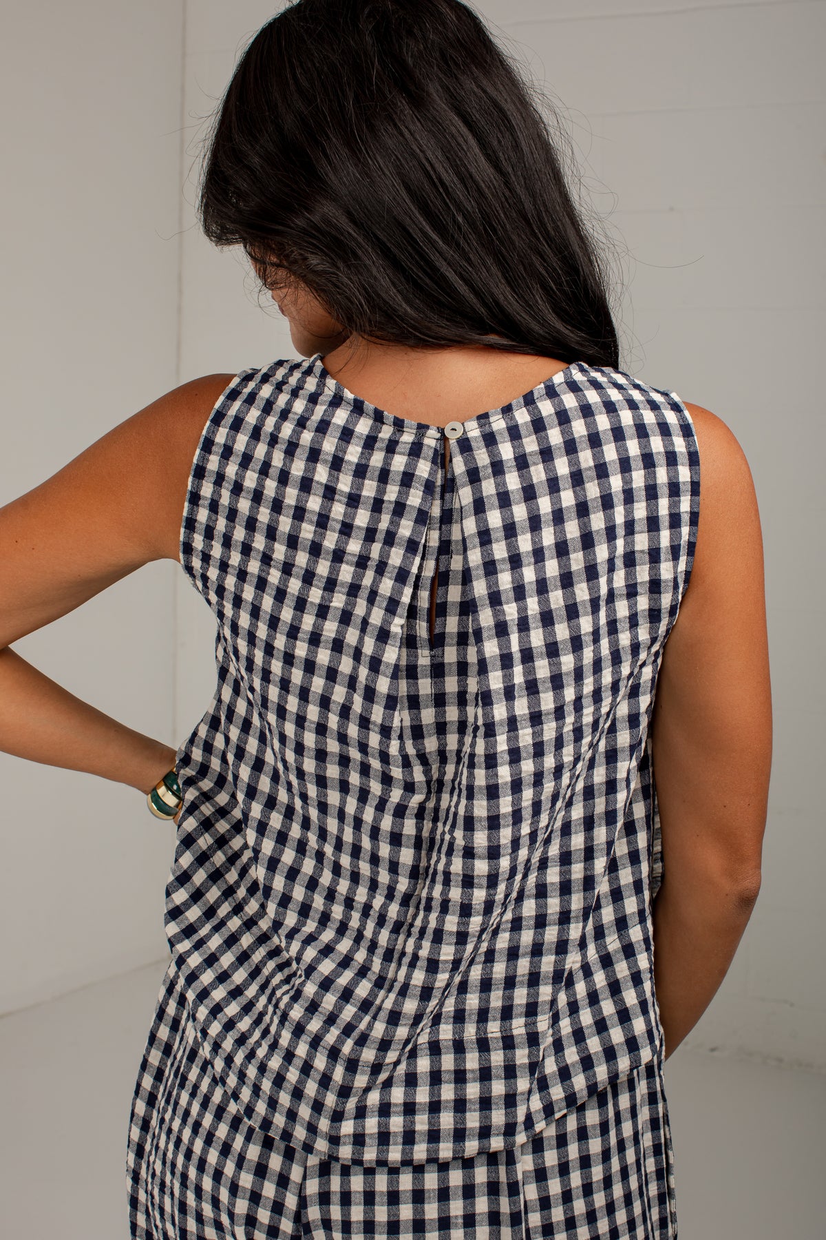 A woman shown from behind wearing a sleeveless navy and white gingham dress with a wrap-style back detail, set against a plain light gray wall.