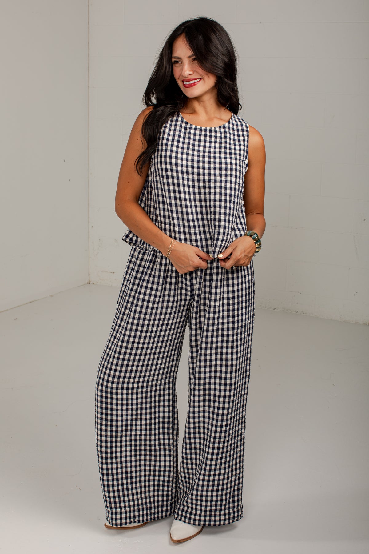 A woman modeling a sleeveless black and white gingham jumpsuit with wide legs, standing in a plain studio setting.