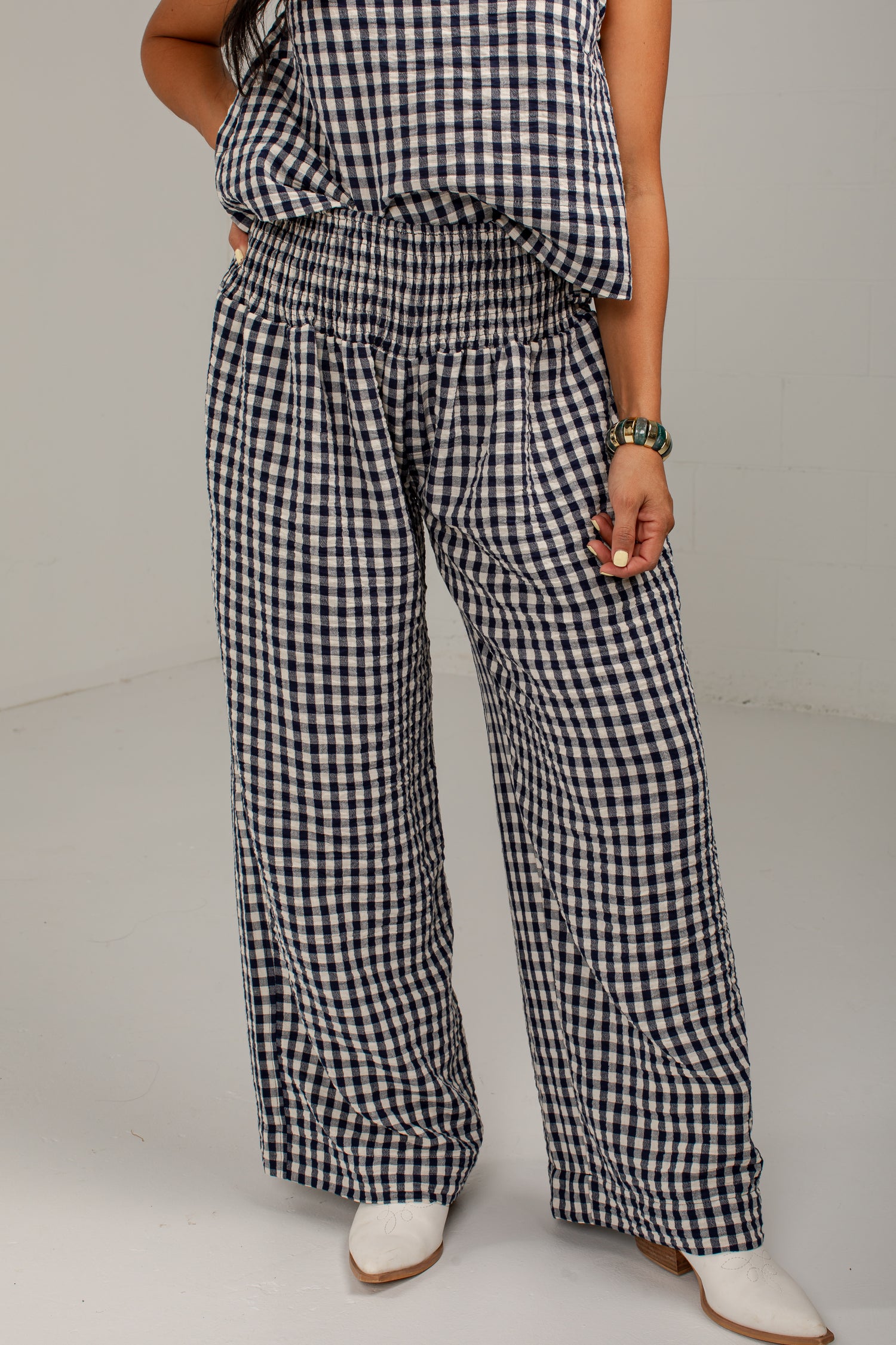 A person wearing loose black-and-white gingham wide-leg pants with an elastic waistband, paired with a matching gingham top and white pointed-toe boots, photographed against a plain light gray studio background.