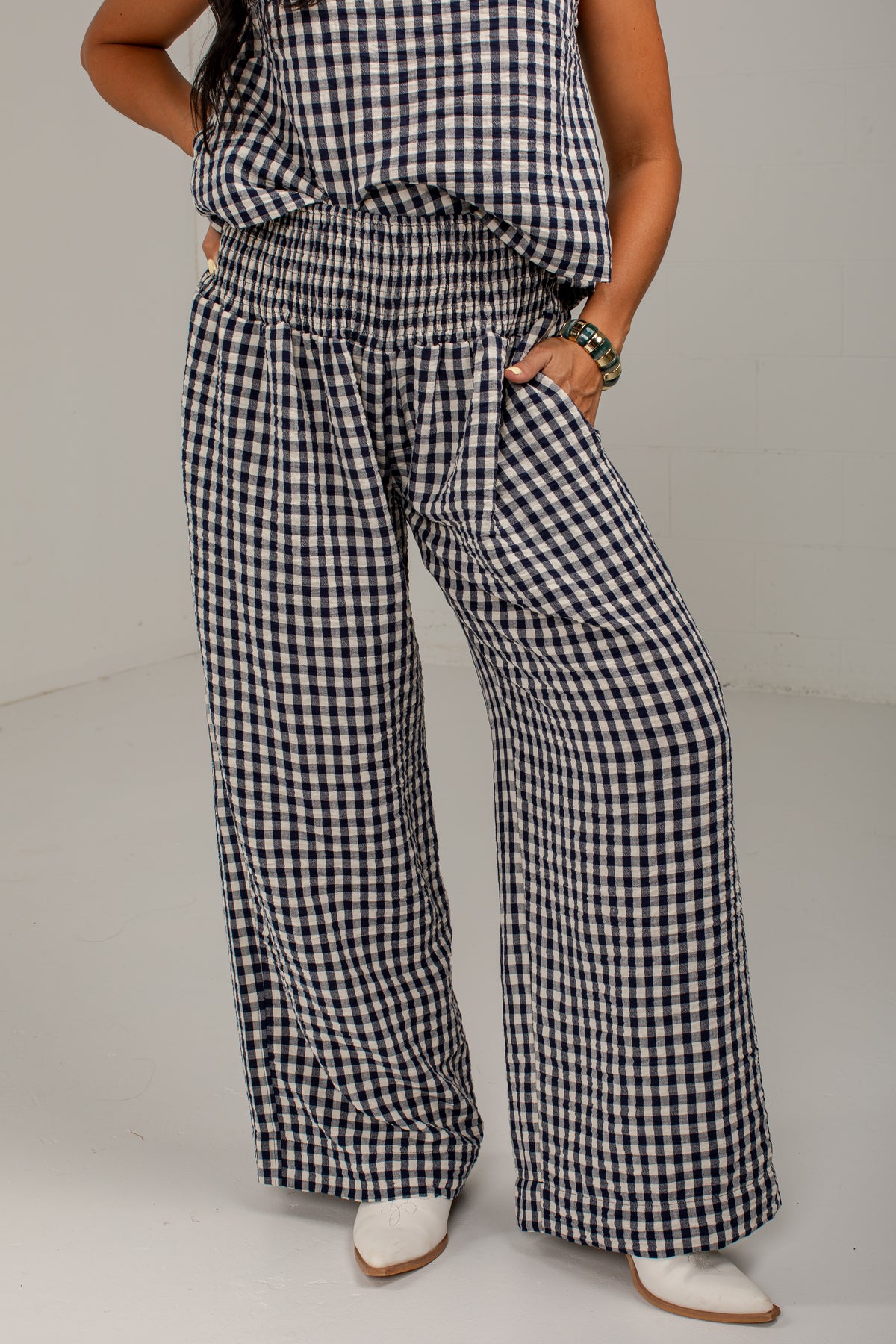 A person wearing loose black-and-white gingham wide-leg pants with a smocked waistband, paired with a matching top and white pointed shoes, photographed against a plain light gray studio background.