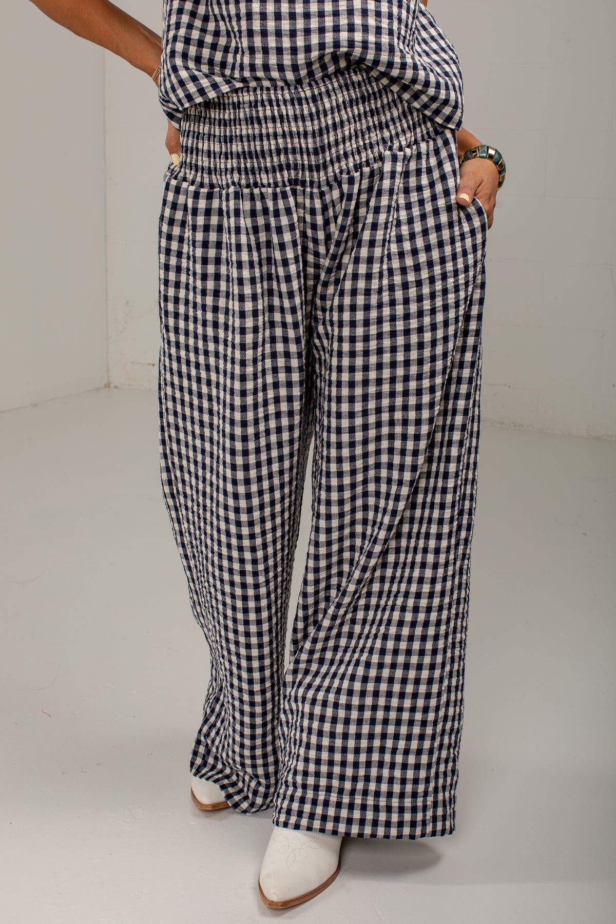 A person wearing loose, wide-leg pants in a black and white gingham check pattern, paired with white pointed-toe boots, standing against a plain light gray studio background.