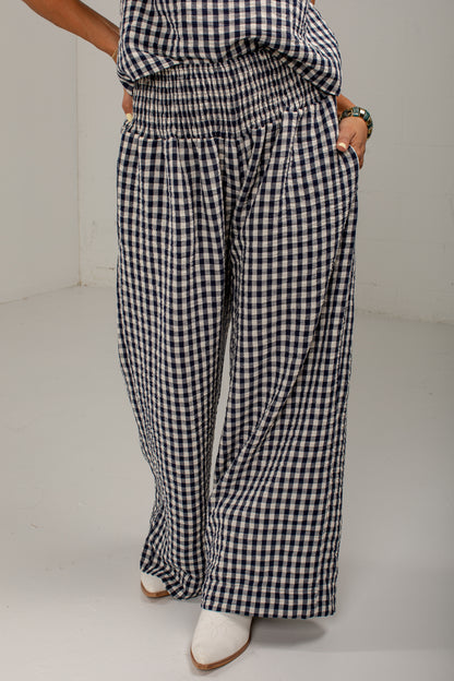A person wearing loose, wide-leg pants in a black and white gingham check pattern, paired with white pointed-toe boots, standing against a plain light gray studio background.