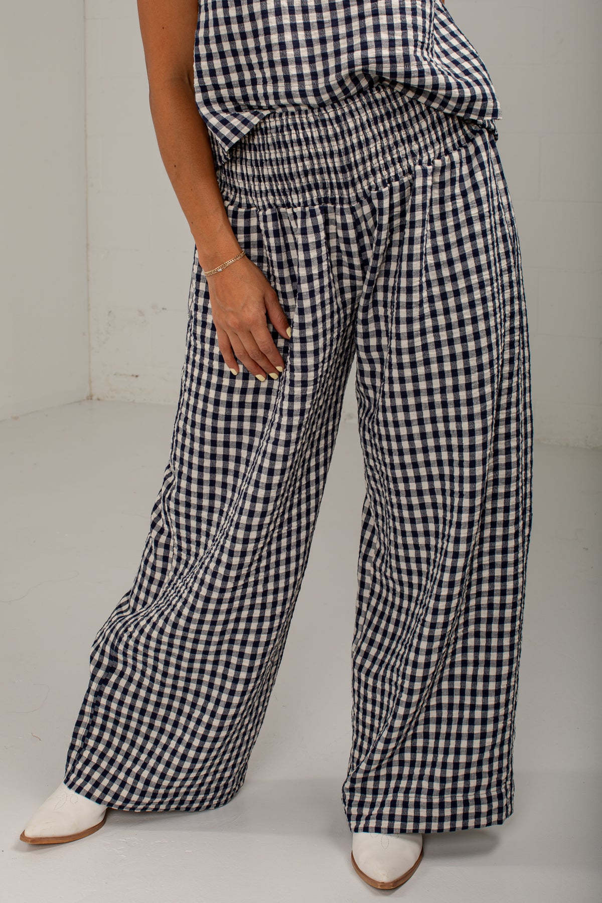 A person wearing loose, wide-leg pants with a black and white gingham check pattern, paired with a matching top and white pointed shoes, photographed indoors against a plain light wall and floor.