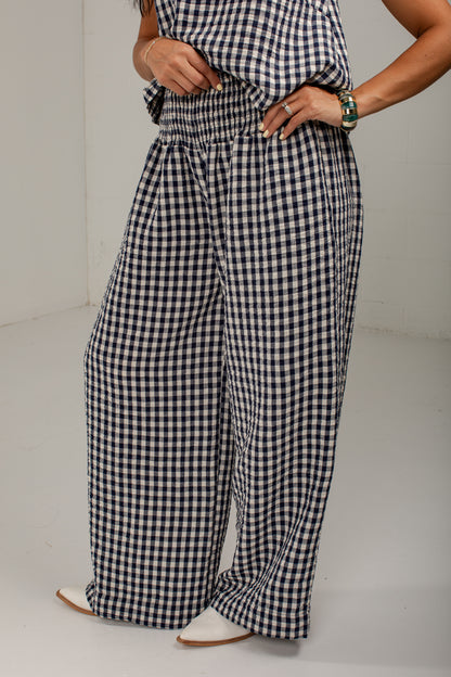 A person wearing loose, wide-leg gingham pants in a black and white check pattern, paired with a matching gingham top and white pointed shoes, photographed in a plain indoor setting.