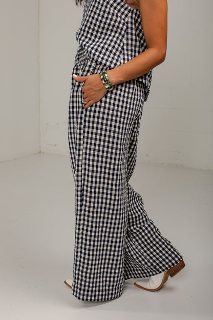 A person wearing a black and white gingham wide-leg jumpsuit, shown from the waist down in a studio setting with a white floor and wall.