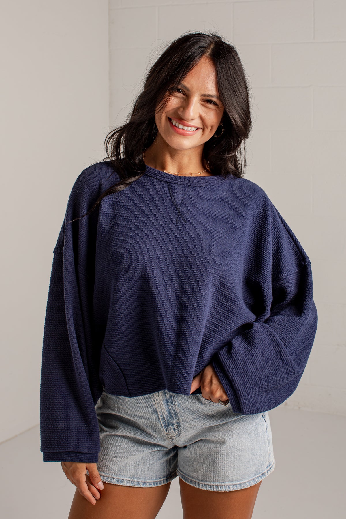 A woman wearing a navy blue textured crewneck sweatshirt and light-wash denim shorts, standing against a light gray brick wall.
