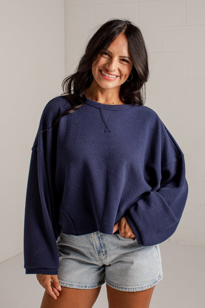 A woman wearing a navy blue textured crewneck sweatshirt and light-wash denim shorts, standing against a light gray brick wall.