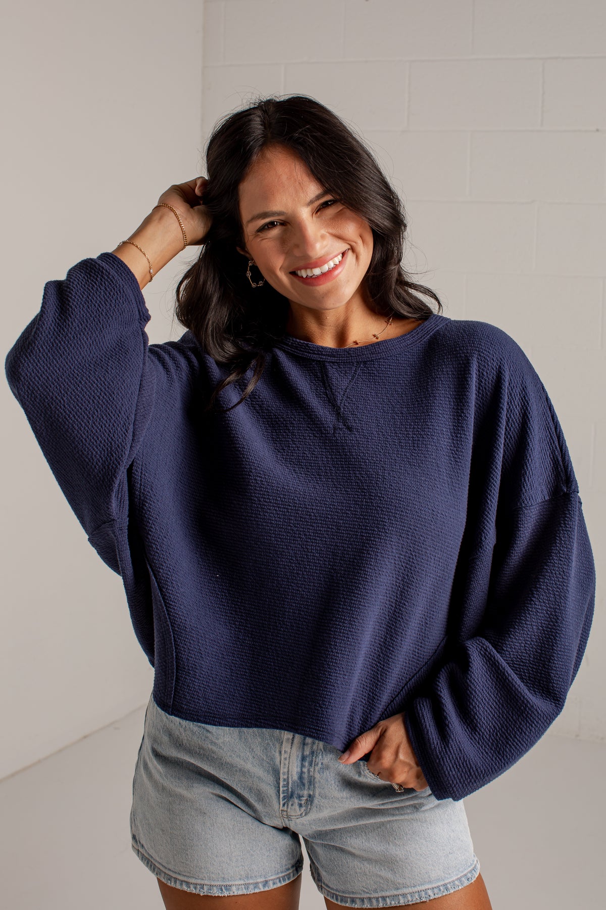 A woman wearing a navy blue textured long-sleeve sweatshirt and light-wash denim shorts, posing in front of a white brick wall.