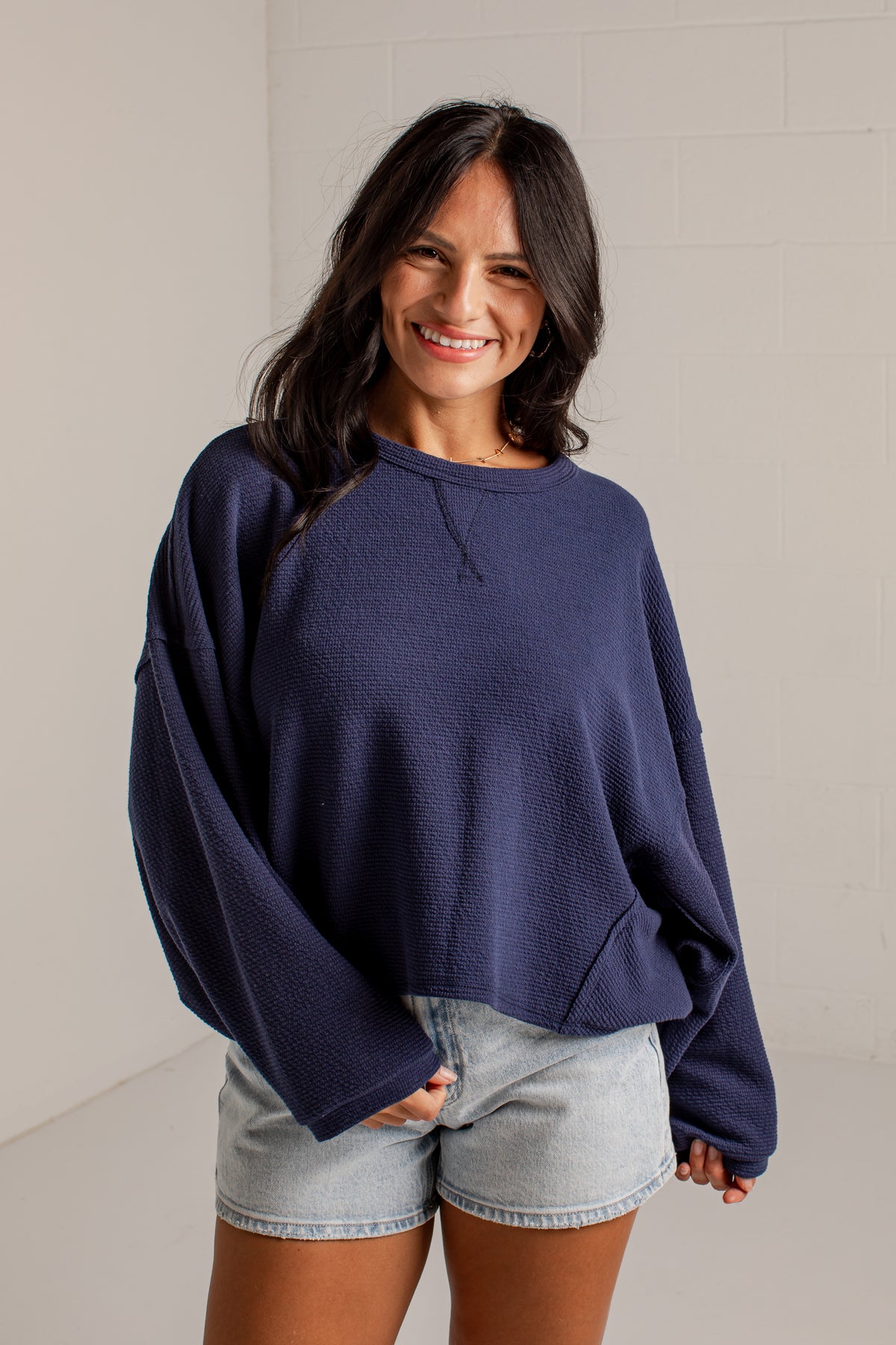 A woman wearing a navy blue oversized crewneck sweatshirt and light-wash denim shorts, standing against a light-colored brick wall.