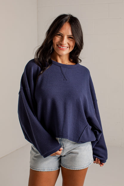 A woman wearing a navy blue oversized crewneck sweatshirt and light-wash denim shorts, standing against a light-colored brick wall.