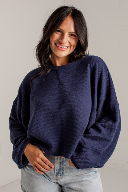 A woman wearing a navy blue textured knit sweater and light-wash jeans, standing against a light gray wall.