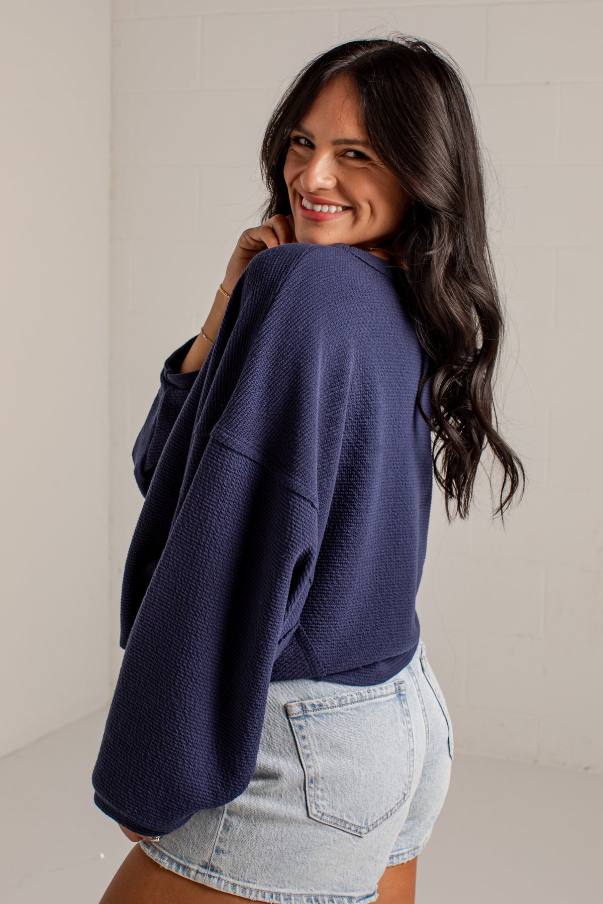 A woman wearing a navy blue waffle-knit cropped sweater and light-wash denim shorts, photographed indoors against a light neutral wall.