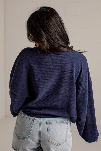 A person viewed from behind wearing a navy blue textured knit sweater and light-wash denim jeans against a plain off-white wall.