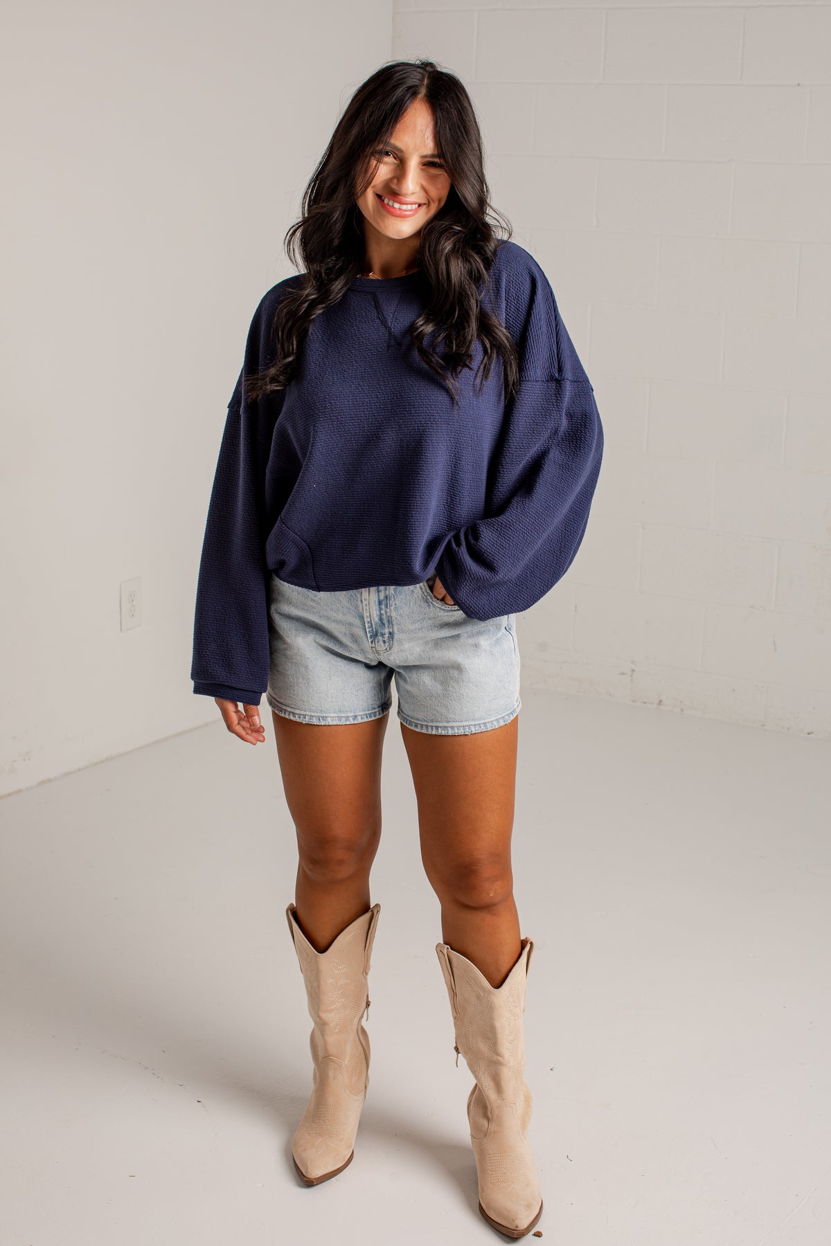A woman modeling a navy blue oversized crewneck sweatshirt, paired with light-wash denim shorts and beige cowboy boots, standing in a bright white studio setting.