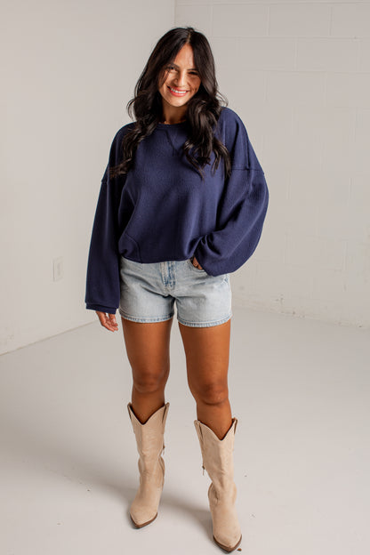 A woman modeling a navy blue oversized crewneck sweatshirt, paired with light-wash denim shorts and beige cowboy boots, standing in a bright white studio setting.