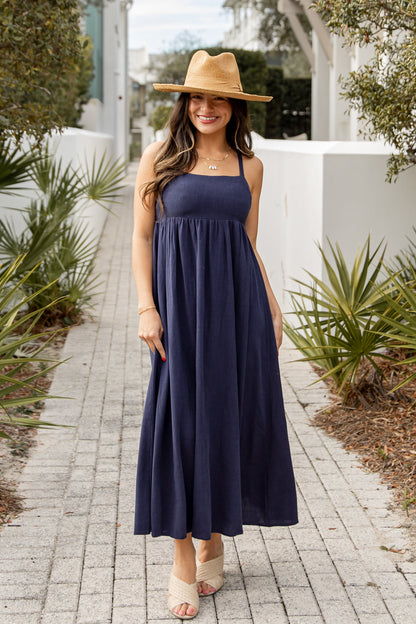 Woman in a navy blue dress and straw hat standing on a paved path with greenery around.