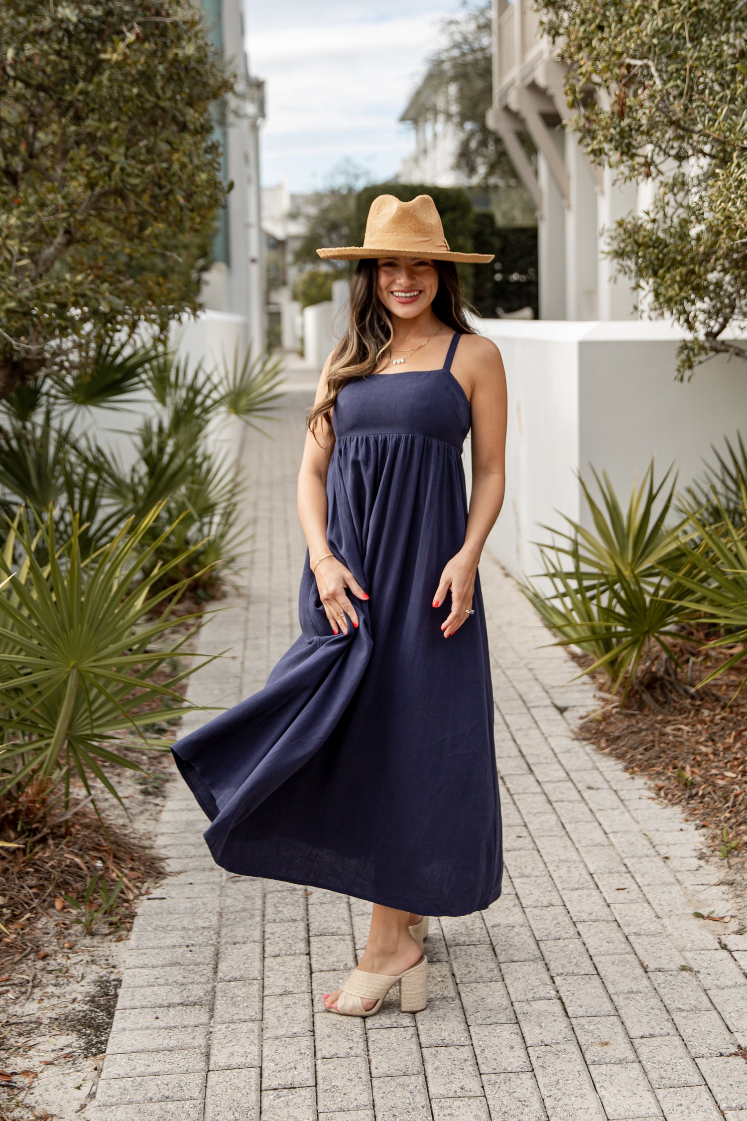 Woman in a navy blue dress and straw hat standing on a paved path with greenery.