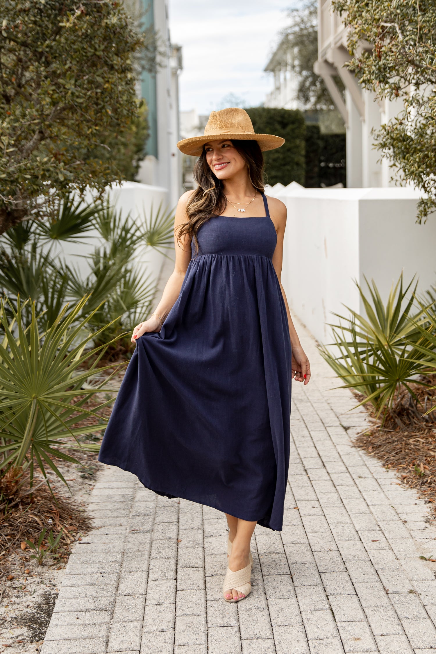 Woman in a navy dress and straw hat walking on a brick path with plants around.