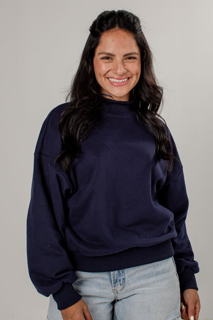 A smiling woman with long dark hair wearing a navy blue mock neck sweatshirt and light wash jeans, standing against a plain light gray studio background.