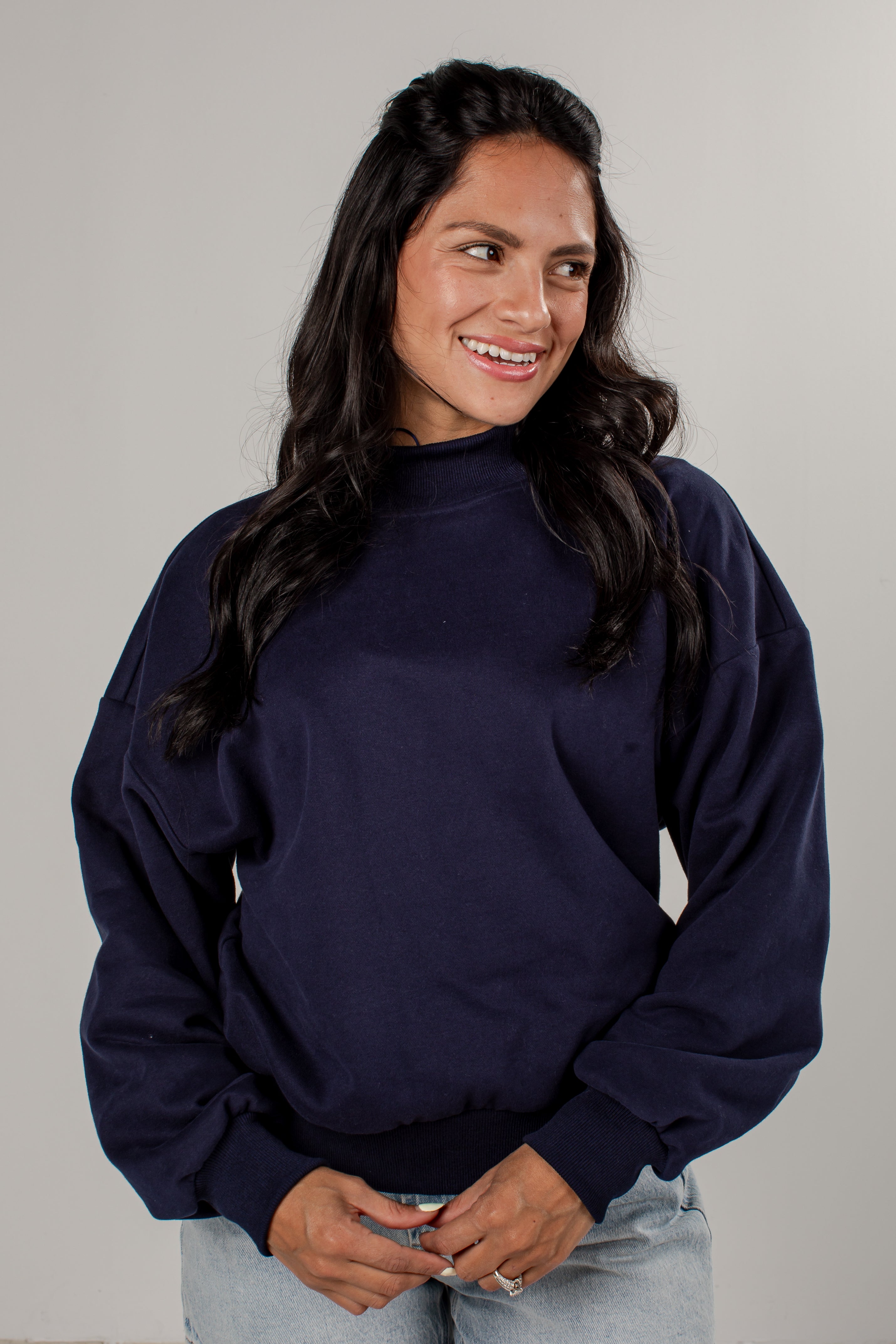 A woman with long dark hair wearing a navy blue mock neck sweatshirt and light wash jeans, standing against a plain light gray studio background.