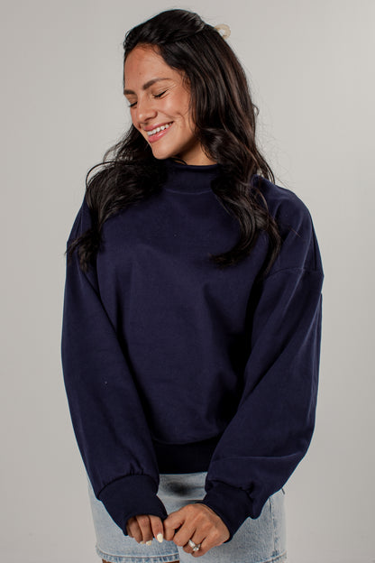 A woman wearing a dark navy blue mockneck sweatshirt and light wash denim jeans, standing against a neutral light gray studio background.