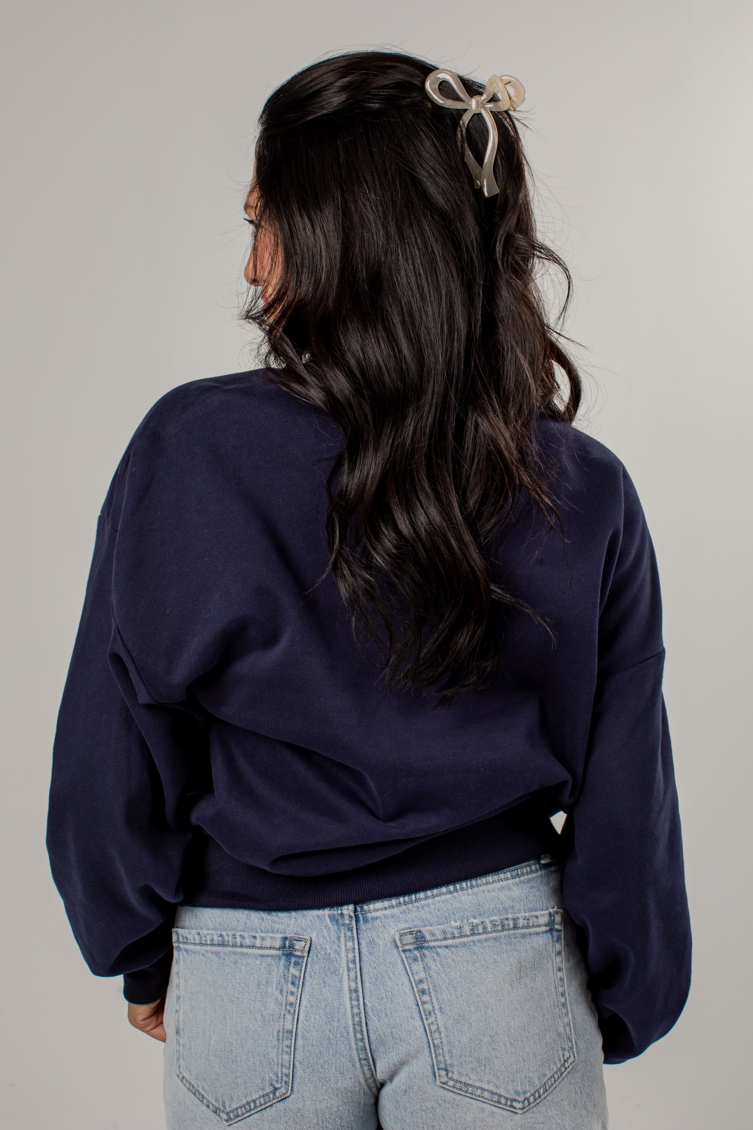 A woman shown from behind wearing a dark navy blue cropped sweatshirt and light wash blue jeans, with a beige bow hair clip in her hair, against a plain light gray background.