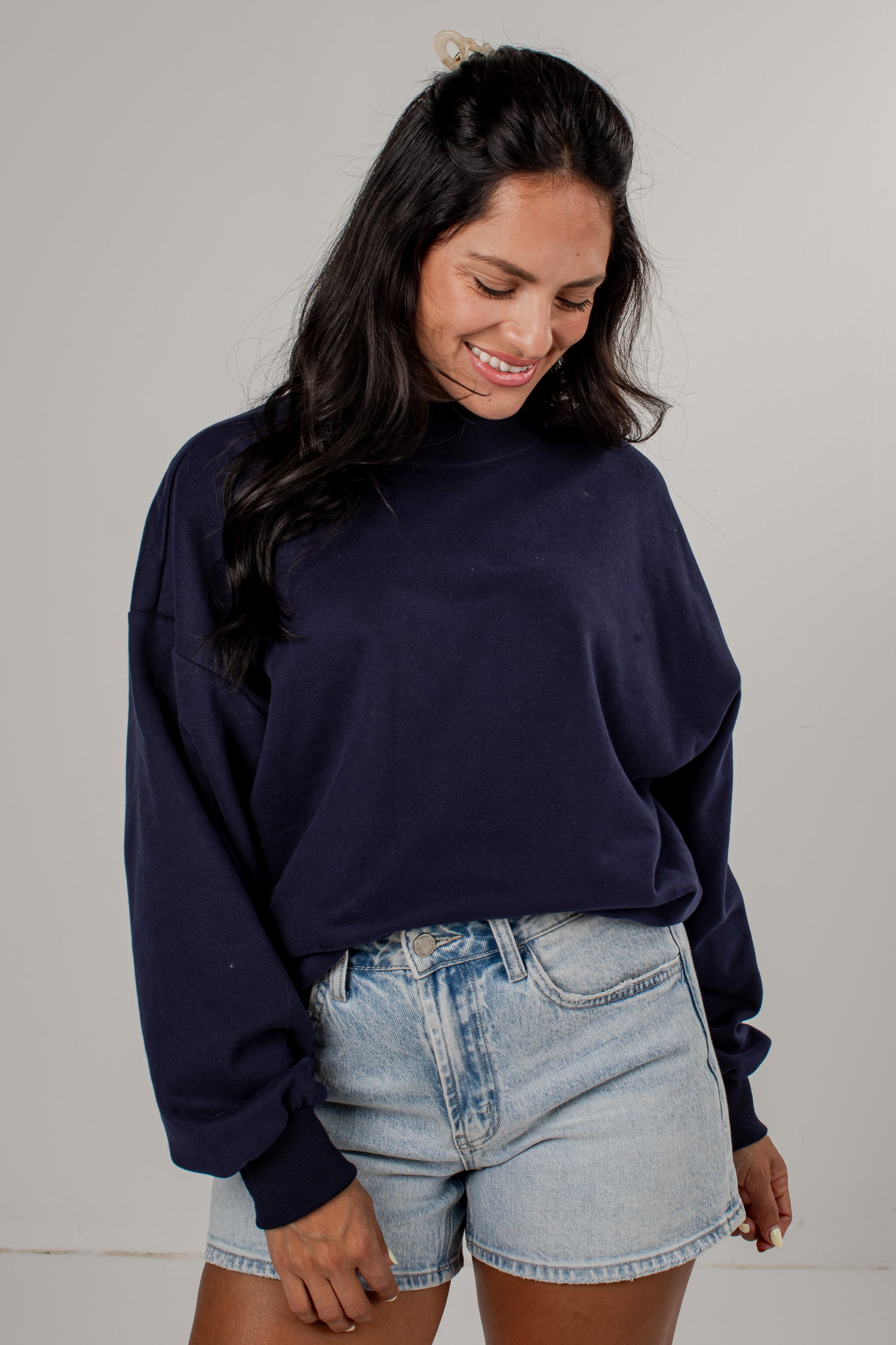 A woman wearing a dark navy blue crewneck sweatshirt and light-wash denim shorts, standing against a plain light gray studio background.