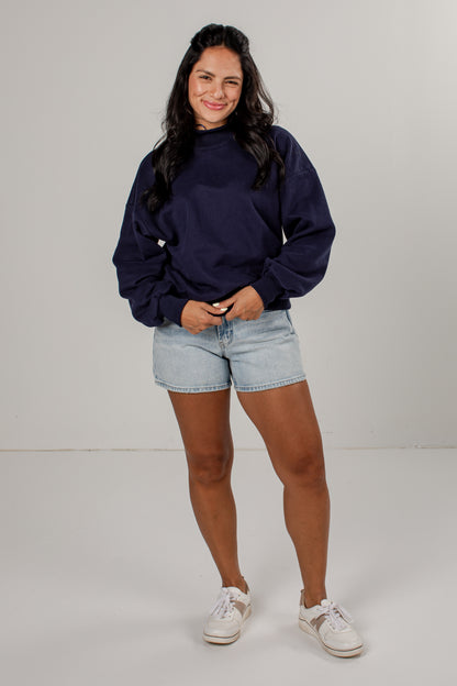 A woman modeling a navy blue sweatshirt with voluminous balloon sleeves, paired with light-wash denim shorts and white sneakers, standing against a neutral light gray studio background.