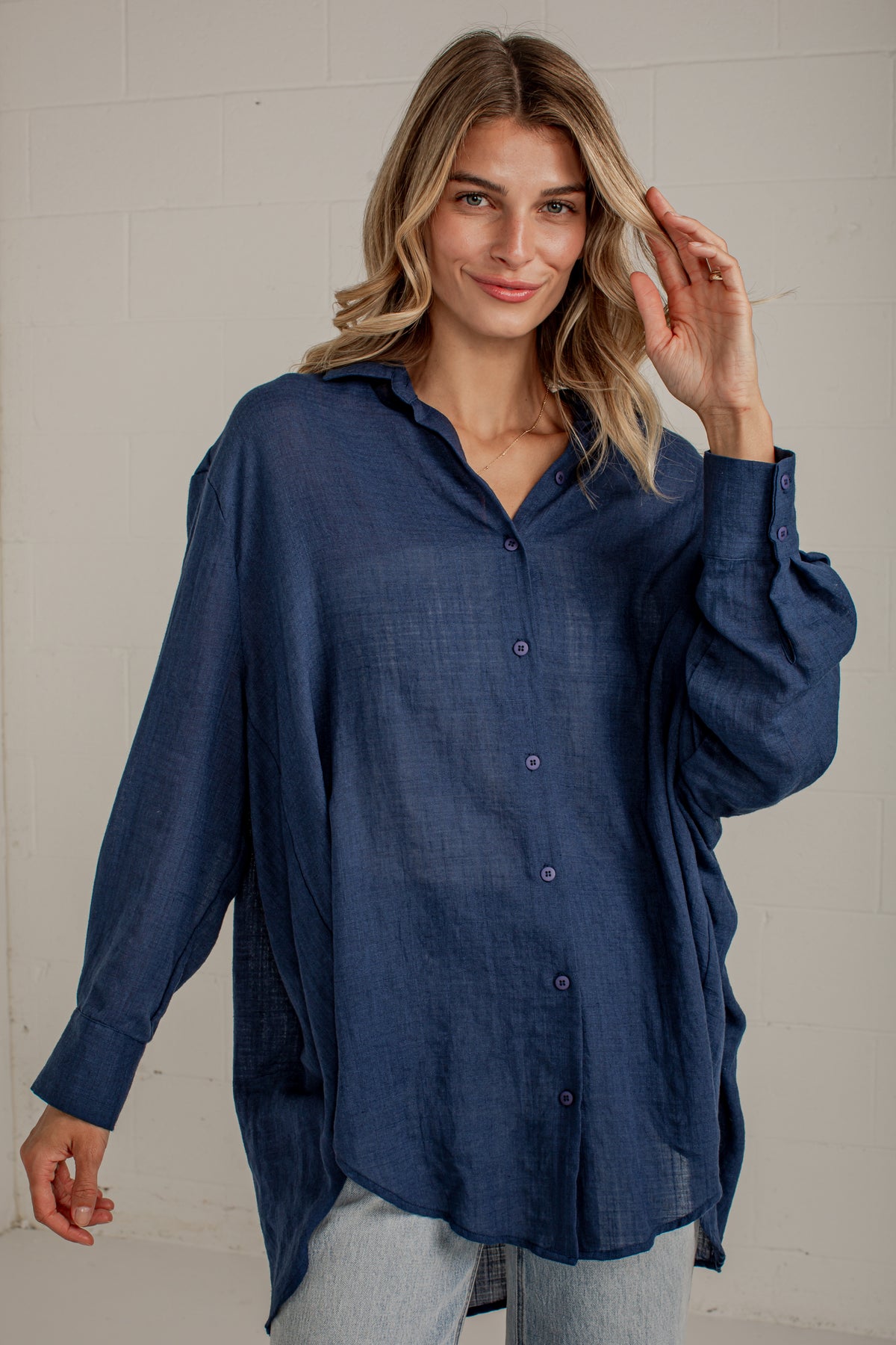 A woman wearing an oversized navy blue button-up shirt with a textured linen-like fabric, paired with light-wash jeans, standing against a white brick wall.