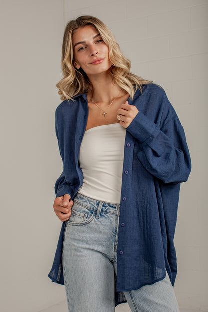 A woman wearing a loose, oversized blue button-up shirt over a white tube top and light-wash blue jeans, standing against a light gray wall.