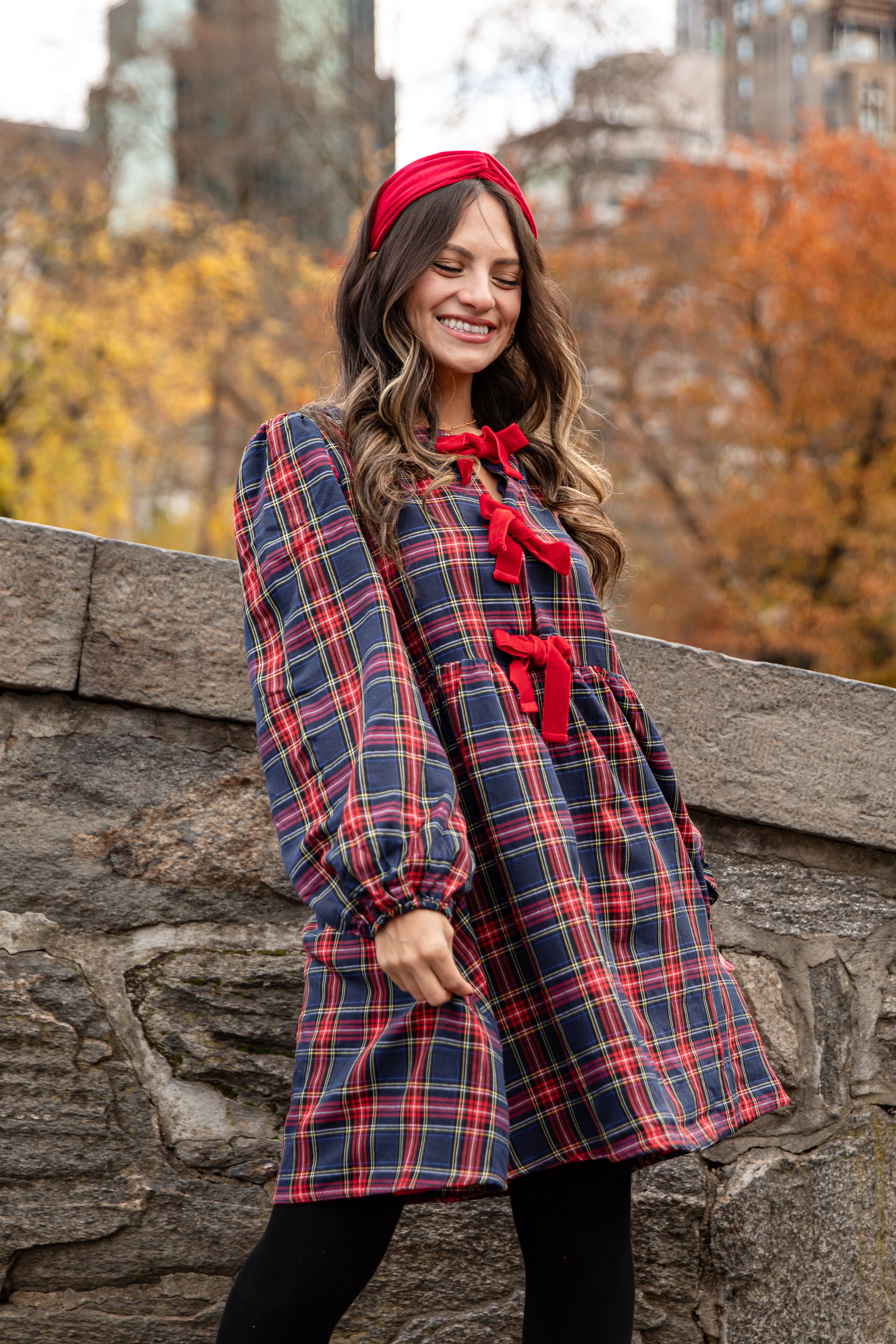 Woman wearing a plaid coat with red accents and a red headband, standing against a stone wall with autumn foliage in the background.