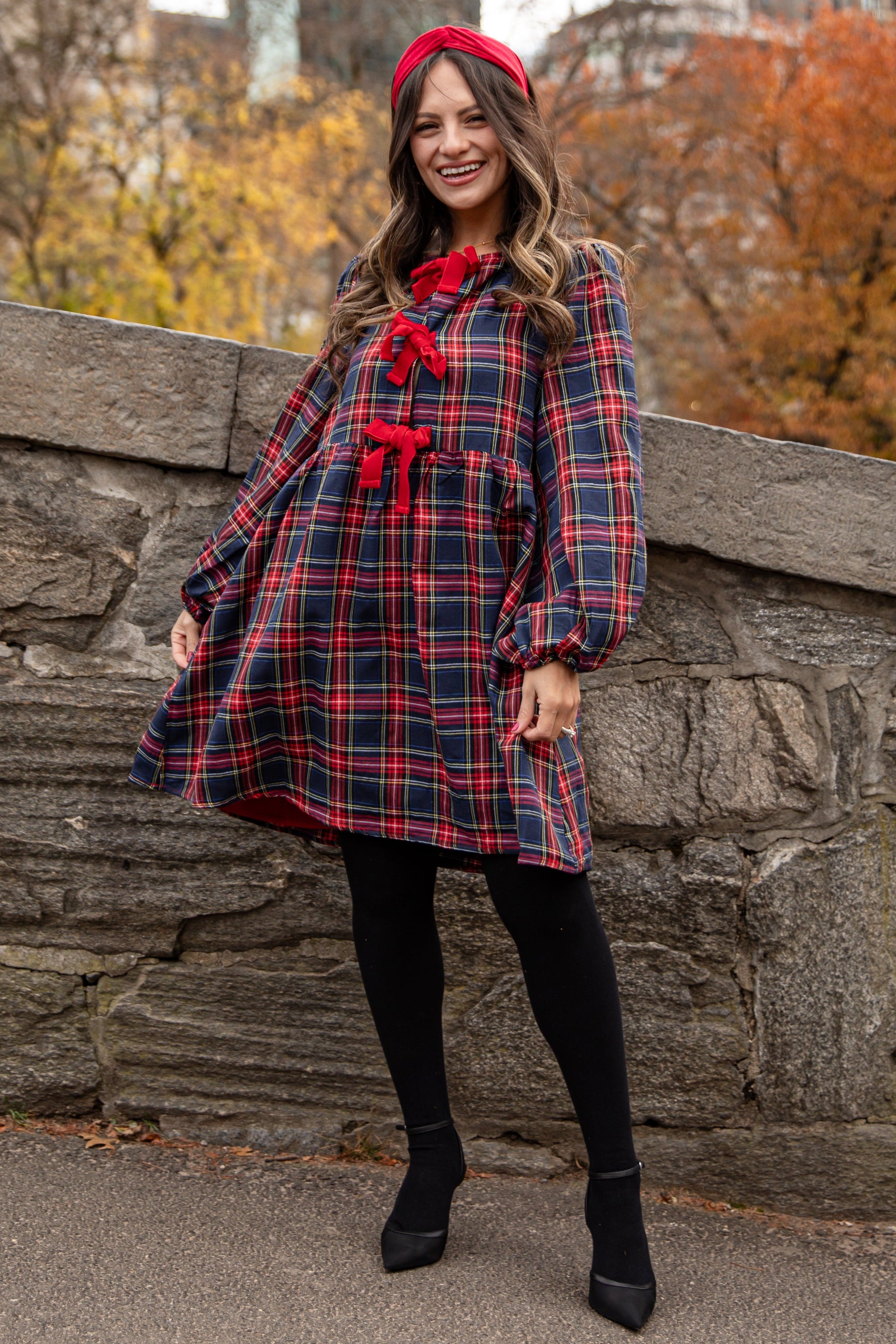 Woman in a plaid dress with red accents standing against a stone wall with autumn trees and city buildings in the background.