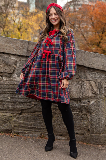 Woman in a plaid dress with red accents standing against a stone wall with autumn trees and city buildings in the background.