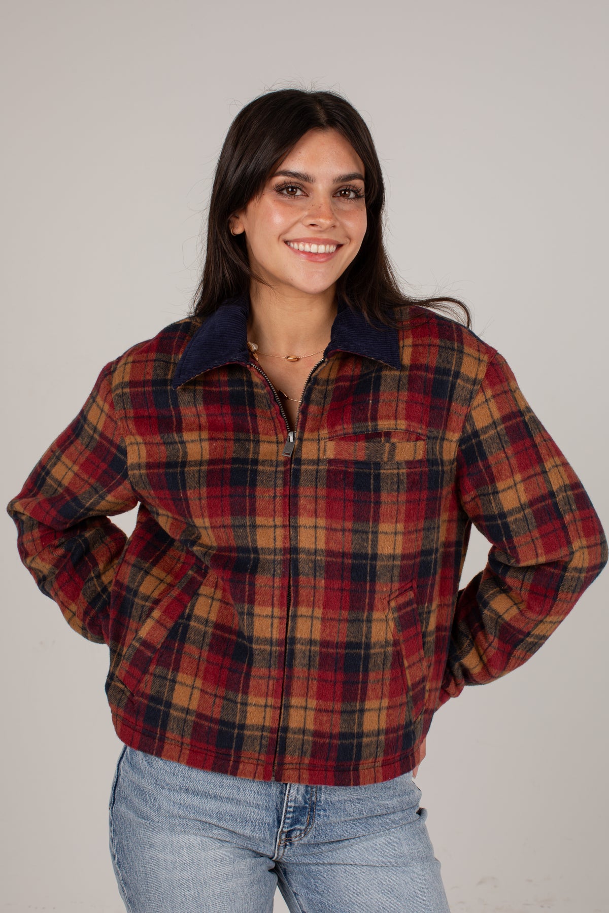 A woman wearing a red and brown plaid zip-up jacket with a dark navy collar, paired with light blue jeans, photographed against a neutral light gray background.