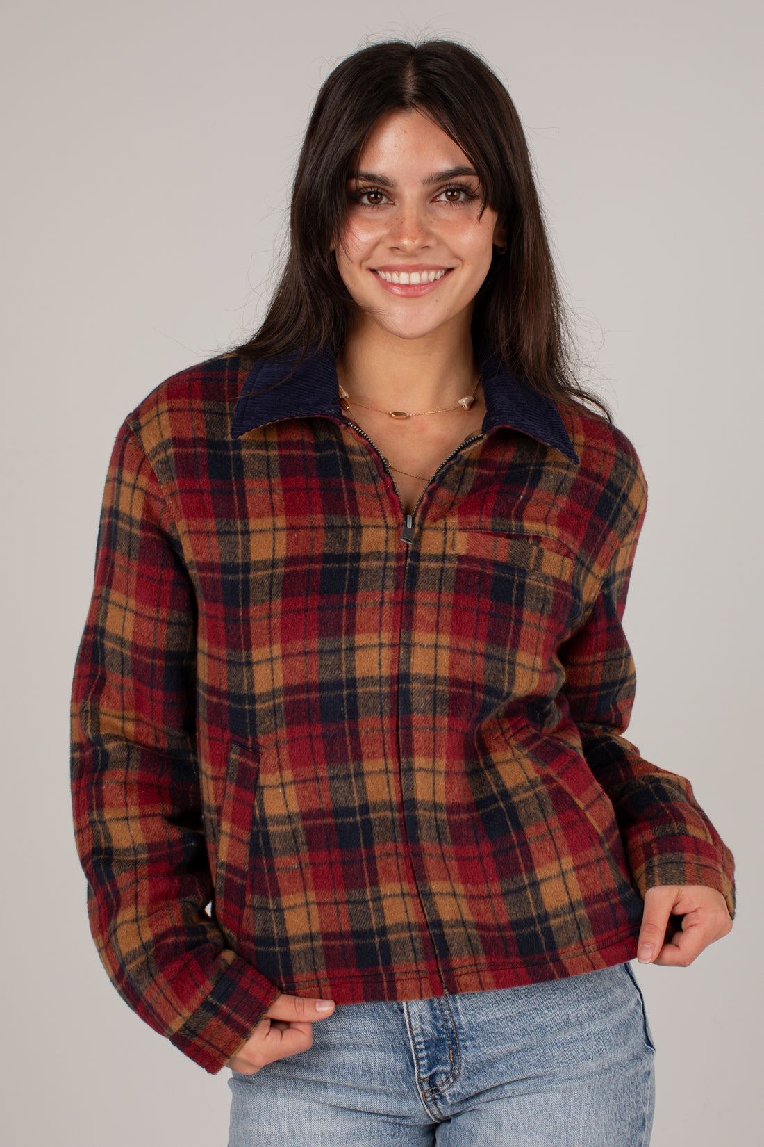 A woman wearing a red, brown, and navy plaid zip-up jacket with a dark navy collar, paired with light blue jeans, against a solid light gray background.