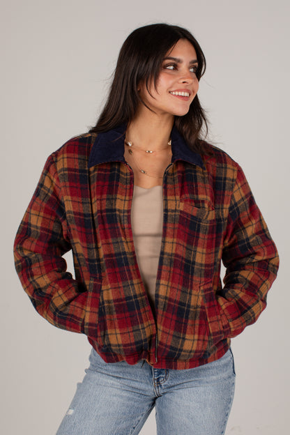 A woman wearing a red, navy, and tan plaid flannel button-up shirt jacket over a beige top and light blue jeans, photographed against a plain light gray studio background.